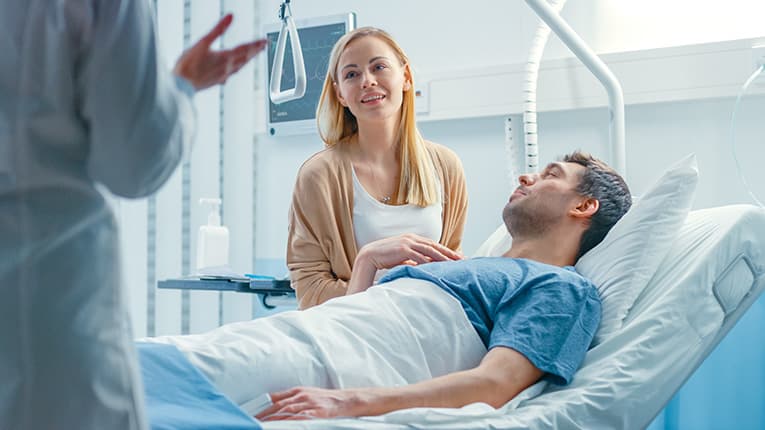a convalescing man in a hospital bed with a caring woman bedside and a doctor advising the woman